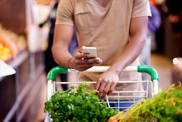 person som stödjer sig över en kundvagn full med mat och tittar i sin smartphone