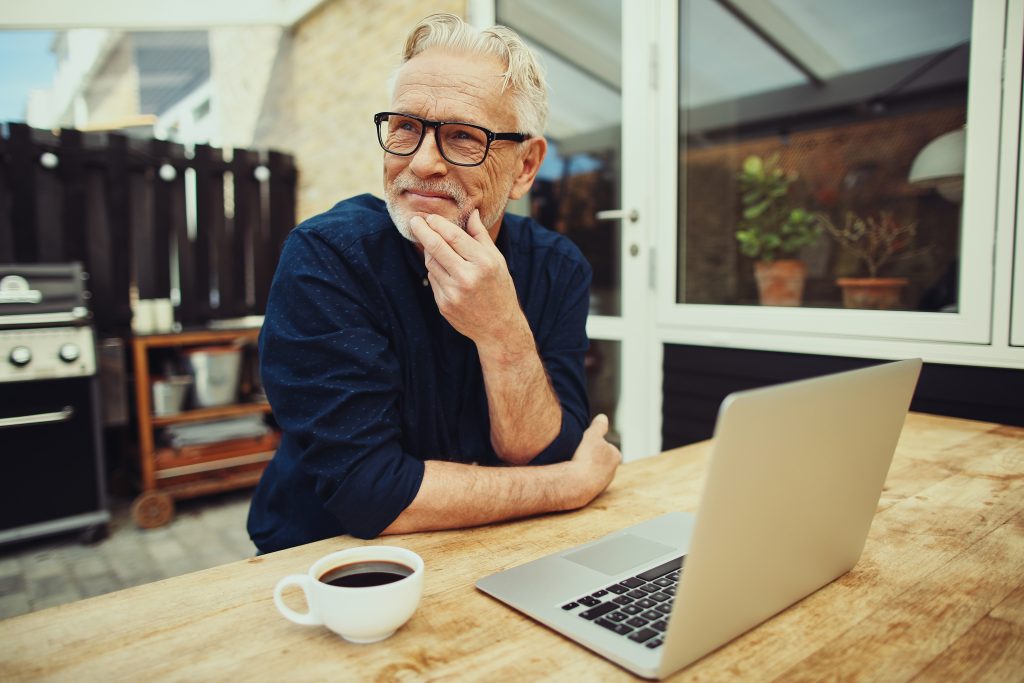 leende äldre man sitter vid bord med en laptop framför sig och en kopp kaffe