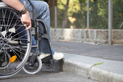 På bilden ses en rullstol vid en trottoarkant. Bilden är beskuren så att man bara ser högra stora och högra lilla hjulet på rullstolen från sidan samt personens jeansklädda ben och högra handen och en bit av underarmen. Rullstolen står framför en trottoarkant.