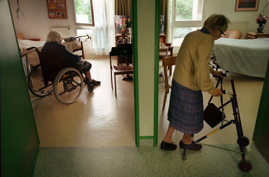 I förgrunden ses en äldre kvinna gå med rollator i en korridor. I bakgrunden ses en annan äldre person sitta i en rullstol