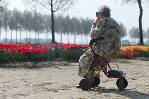 en äldre kvinna sitter på sin rollator. Hon befijner sig i en park, framför henne finns en rabatt med röda och gula blommor och längre bort en rad med träd. Vi ser henne snett bakifrån och hon tittar ut över rabattera. 