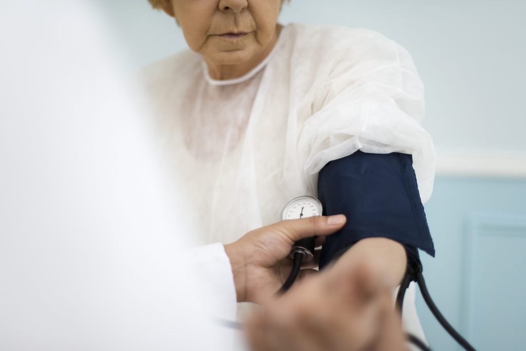Bilden visar hur en patient, en äldre kvinna klädd i vit blus får sitt blodtryck mätt. en blå blodtrycksmanschett sitter på hennes vänstra arn