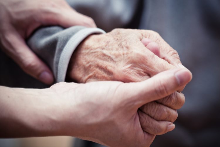 en rynkig hand som tillhör en äldre person hålls om av ett par yngre händer
