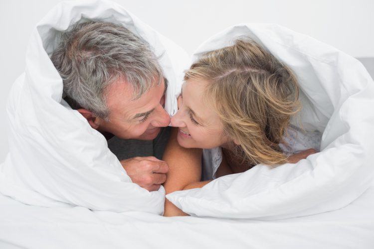 medelålder par ligger i sängen invirade i vita täcke så att bara deras huvuden sticker fram. De ler och gnuggar sina näsor mot varandra