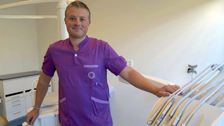 På bilden ses Björn Gjelvold iklädd lila tandläkarskjorta. Han står bredvid ett ställ med tandläkarborrar med flera instrument.