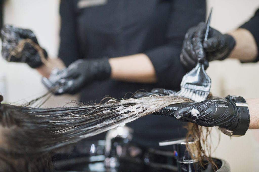Bilden visar hur en frisör färgar håret på en person. I detalj ses en hårslinga som hålls upp av en handsbeklädd hand. Den andra handen håller i en pensel med vilken frisörer applicerar hårfärg på hårslingan 