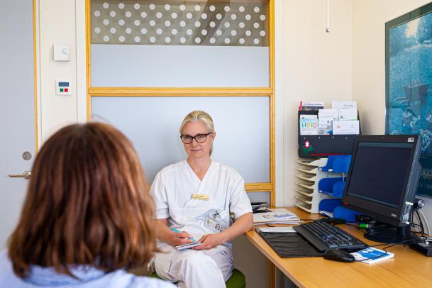 På bilden ses Åsa Petersén klädd i vita läkarkläder. Hon sitter på sitter tjänsterum med ett anteckningsblock i knät och pratar med en persons som på bilden syns bakifrån