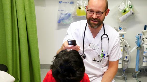 på bilden ses läkaren Kees-Jan Pronk klädd i vita sjukhuskläder. Han sitter på en pall i ett undersökningsrum med ett stetoskop runt halsen. Han håller i något som ser ut som en ficklampa som lyser. Framför honom sitter ett barn. Vi ser barnets huvud snett uppifrån. 