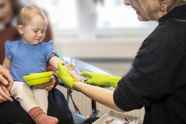 På bilden ses ett litet barn i ettårsåldern. Barnet sitter i knät på en vuxen. En sköterska med limegröna handskar på händern tar en blodprov på barnet. Barnet ser tveksamt ut men gråter inte