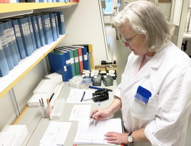 Carina Törn är klädd i vit labbrock. Hon står vid en labbänk på vilken det står pärmar, provrörsställ och en del papper. Hon ser ut att fylla i ett labbprotokoll