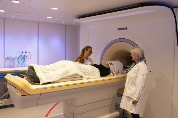 Pia Sundgren och Linda Wennberg står på var sin sida om en patient som ligger på en släde som är på väg att skjutas in i magnetkamerans tunnel
