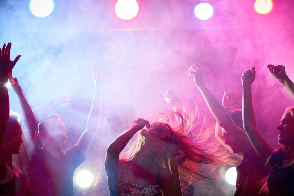 Ungdomar som dansar på en klubb. I bildens mitt ses en ung kvinna med lång hår. I bakgrunden flera andra personer som dansar. Allt ses i lilafärgat nattklubbsljus