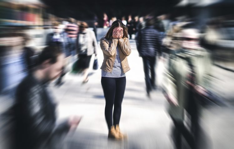 Ung kvinna står och håller händerna för ansiktet. Hon omges av en massa andra människor som passerar henne i olika riktningar. I bilden är hon i fokus med de passerande människorna är suddiga. Bilden illustrerar en panikångestsiruation