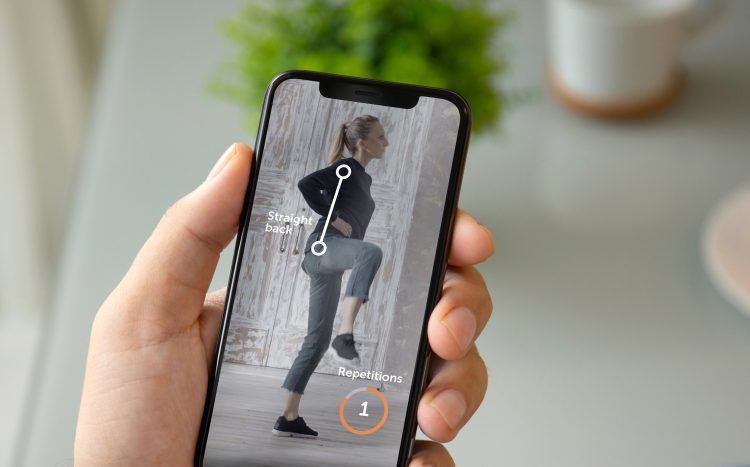 På bilden ses en hand som håller i en smartphone. På skärmen ser man en kvinna göra en övning och stödillustrationer i bilden visar vad som är rätt hållning