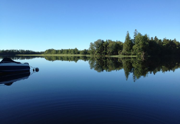 en sommarvy av en sjö med spegelblankt vatten och blå himmel på motsatta stranden ses skog och till vänster i bild ses en ankrad motorbåt