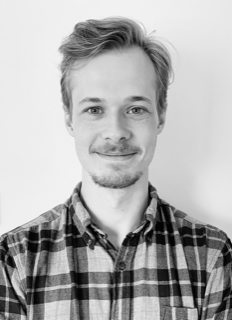 ett svartvitt porträttfoto av Filip Bäckström. Han har mörkblont kortklippt hår, hakskägg, mustasch och är klädd i en rutig skjorta