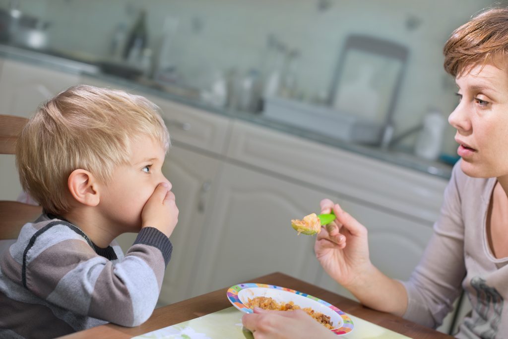 Scenen utspelar sig i ett kök. En liten pojke, 2-3 år, och hans mamma sitter vid matbordet. Pojken som är till vänster i bilden håller för munnen med högra handen samtidigt som hans mamma håller upp en sked med mat framför hans mun. DEn andra handen håller ho om en liten barntallrik full med mat