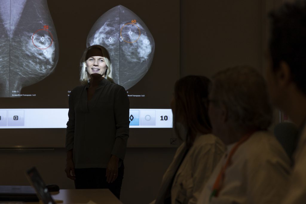 På bilden ser man Sophia Zackrisson när hon föreläser. Hon står framför en skärm på vilken man kan se mammografibilder av bröst. Rummet är nersläckt och hon sys bara i siluett förutom hennes ansikte som är upplyst av ljuset från projektorn. I förgrunden ser man ett par av åhörarna, två kvinnor som ser ut att vara klädda i vita sjukhusrockar.