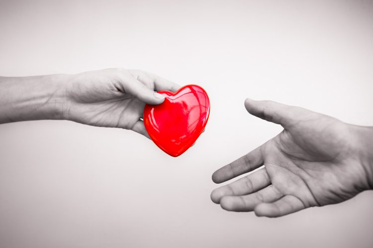på bilden ses två händer där den ena håller i ett rött plasthjärta och räcker det över till den andra handen. Förutom det röda hjärtat är bilden i svartvitt