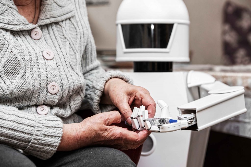 En äldre kvinna klädd i grå kofta tar emot tabletter från en robot