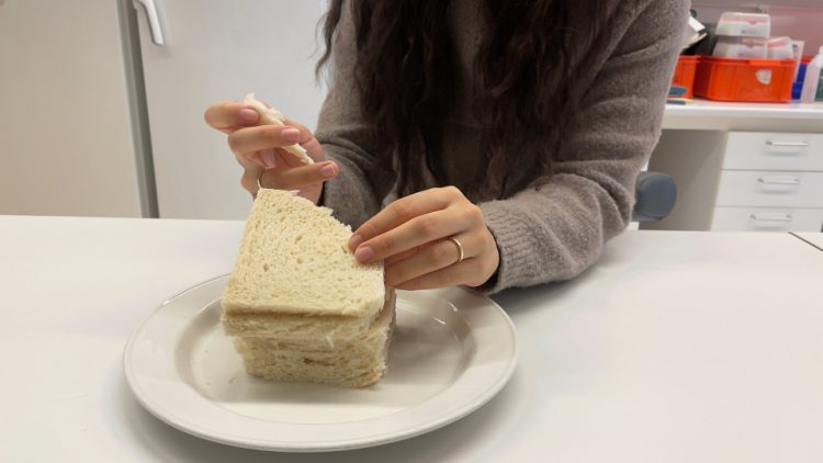 vid tallrik med 5 vita brödskivor staplade på varandra. Kvinnohänder bryter av en bit av brödet. Man ser även kvinnans bål, hon är klädd i grå stickad tröja, och har långt mörkt hår som faller ner över bröstet