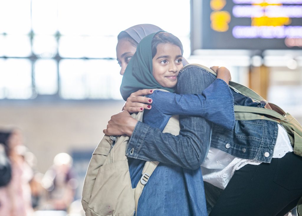 muslims mamma iklädd jeansjacka, byxor och huvudduk kramar om sin lilla dotter, även hon klädd i jeansklänning och huvudduk. Scenen utspelar sig på en flygplats