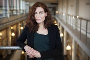 Medelålders kvinna med långt kastanjebrunt hår, klädd i svart kavaj och en mörk topp. Hon lutar sig med ena armbågen mot ett räck. Bakom interiör från diabetescentrum i Malmö