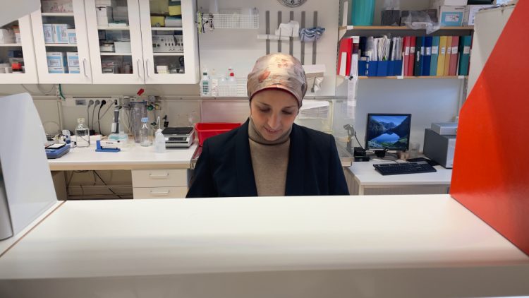 yngre kvinna sitter bakm en bänk på ett labb. Bakom henne syns diverse labbutrustning och pärmar på hyllor. Hon bär huvudduk och är klädd i mörk kavaj och en beige polotröja