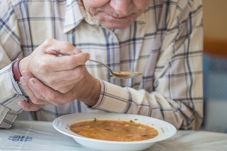 äldre man äter soppa. Håller en sked med soppa på i ena handen och håller med den andra hand om handleden för att hindra skakningar