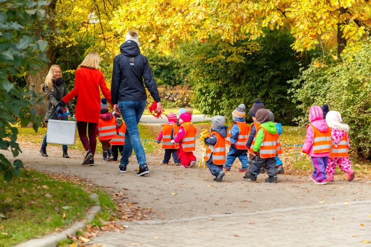 En grupp små förskolebarn på promenad i en park. De har orangea reflexvästar på sig. tillsamman med dem går två personal, två kvinnor. Den ena klädd i röd kappa och brua byxor, den andra i jeans och mörkblå jacka.
