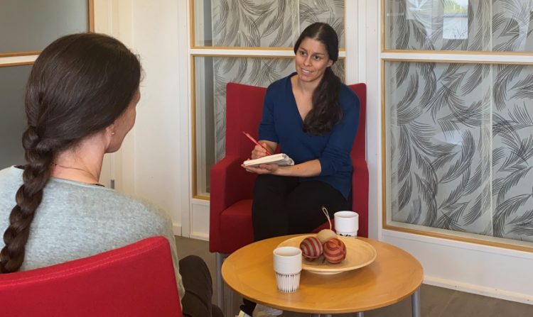 Två kvinnor sitter mitt emot varandra i varsin röd fåtölj med ett runt litet soffbord mellan sig. På bordet står två kaffekoppar och en skål med tre bollar i. Kvinnan till vänster i bild ses bakifrån. Hon har låg mörkt hår uppsatt i en fläta. Hon bär en grå tröja. Kvinnan mitt emot ses framifrån. Hon håller i ett block och en penna. Även hon har lång mörkt hår ihopsatt i en hästsvans och hön är klädd i mörka byxor och en mörkblå skjorta.