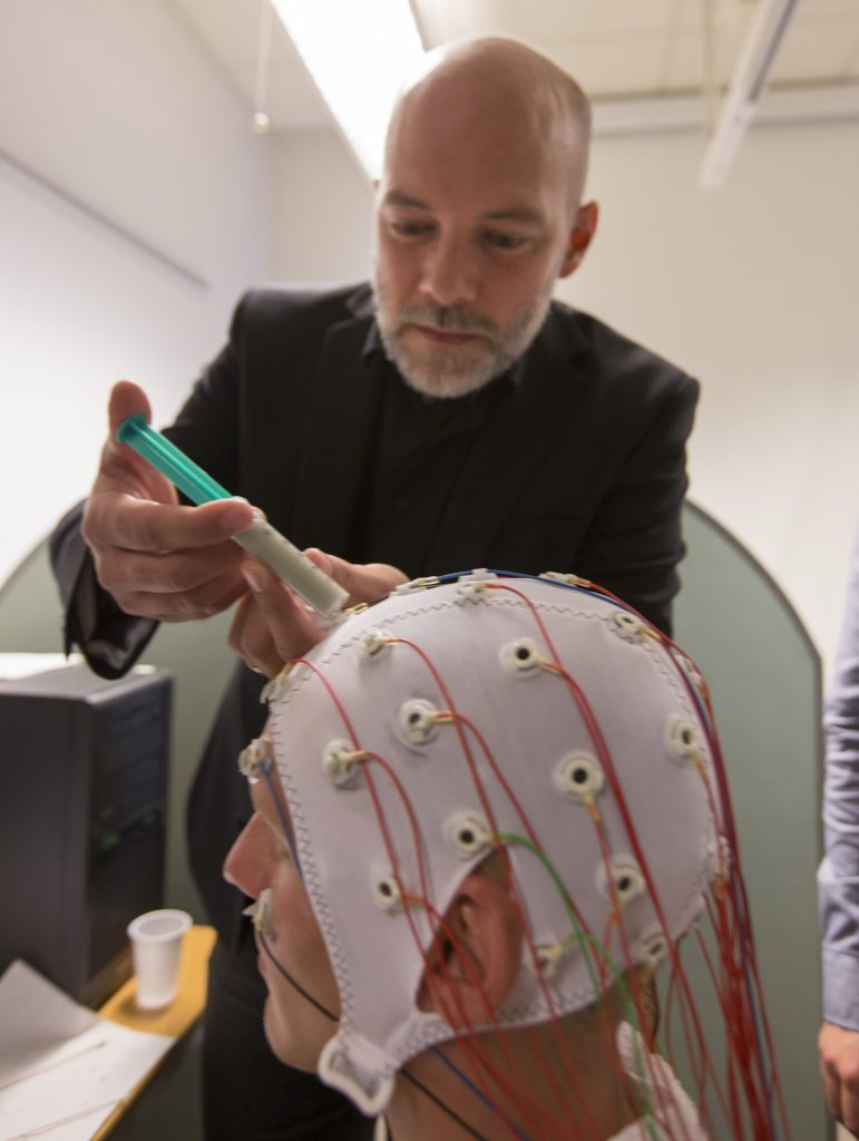 En man sprutar in något vid huvudet på en försöksperson. Försökspersonen har en hätta på huvudet som är full av elektroder. En ung man tittar på..