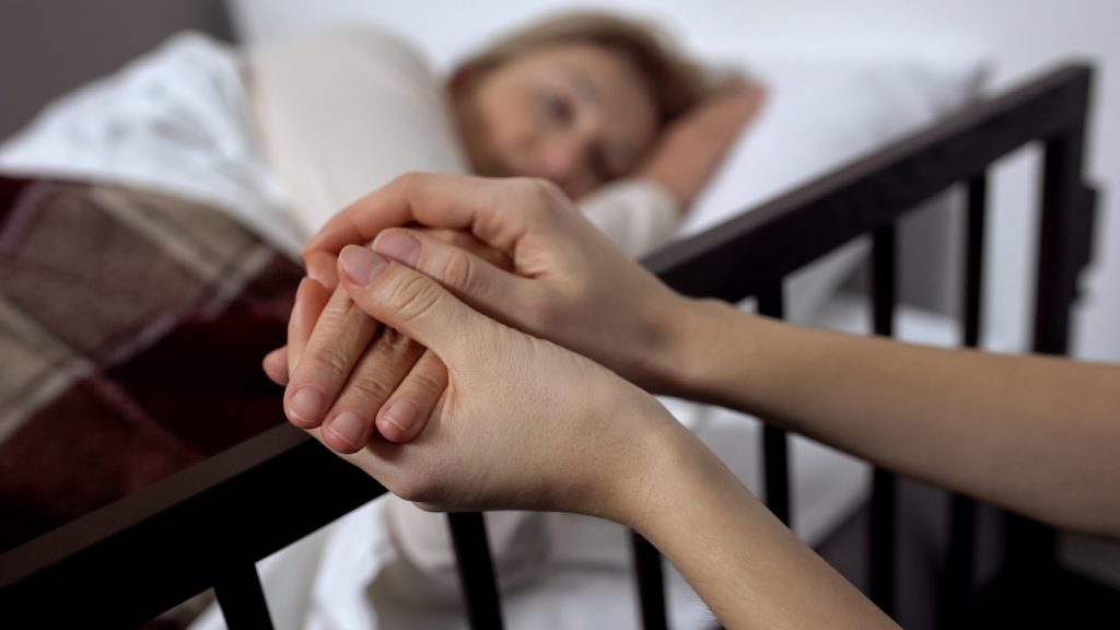 medelålders kvinna ligger i en säng och ett par yngre händer håller hennes ena hand