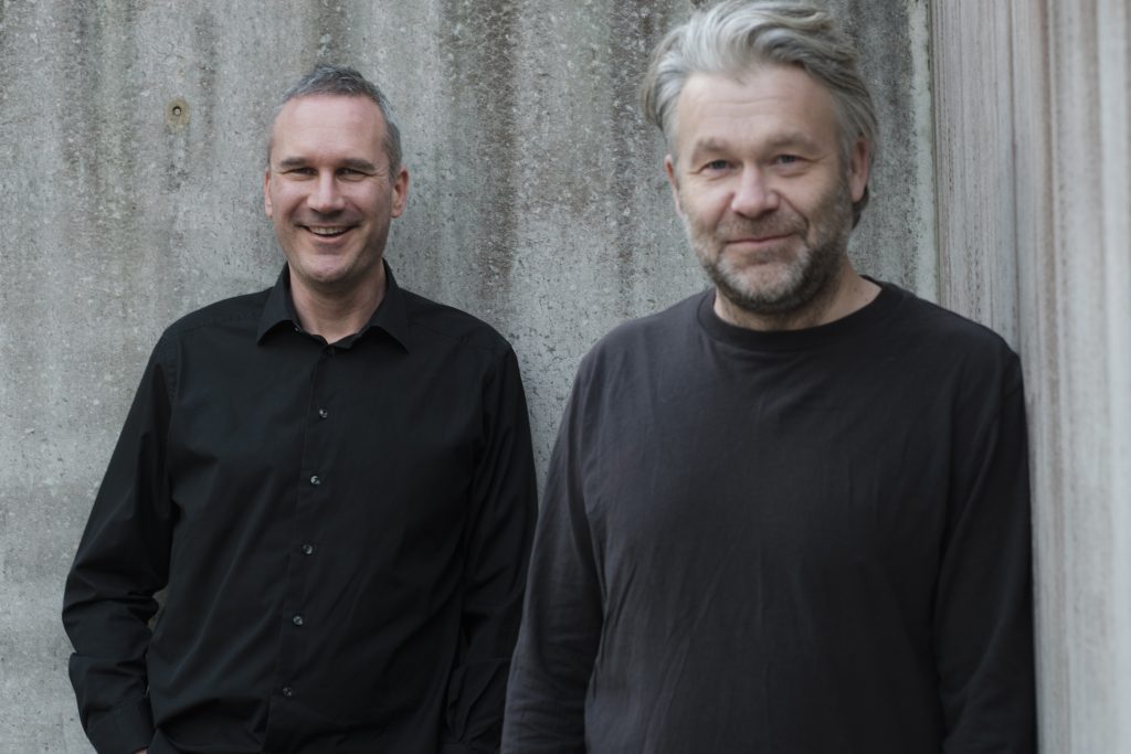 Två män i svart skjorta respektive svart tröja mot grå vägg. 