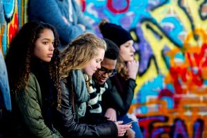 Fyra tonåringar sitter med mobiler framför grafittivägg.