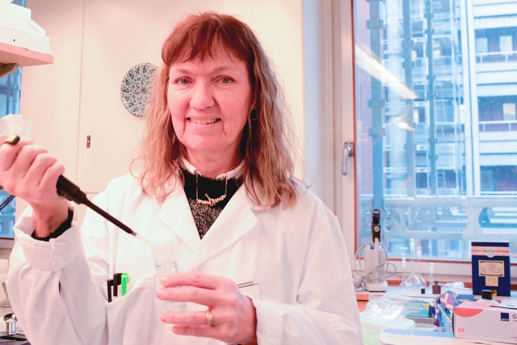 Kvinna i vit labbrock håller en pipett i ena handen och ett plaströr i den andra