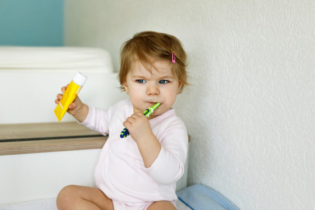 bäbis sitter i badrummet och biter på en tandborste samtidigt håller hon en tandkrämstub i andra handen