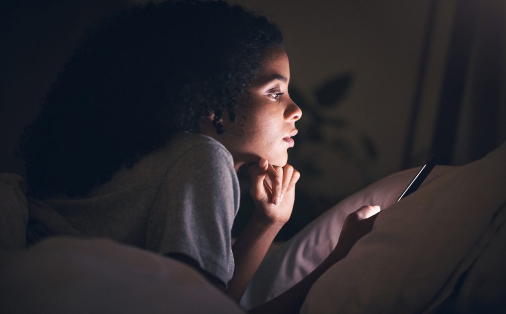 ung kvinna ligger på sängen i mörkt rum och läser på sin smartphone