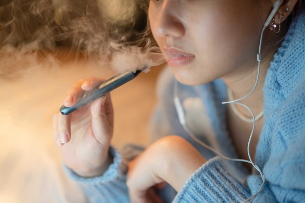 närbild på ung kvinna med hörlurar i öronen och en e-cigarett i handen. Det kommer rök ur hennes mun