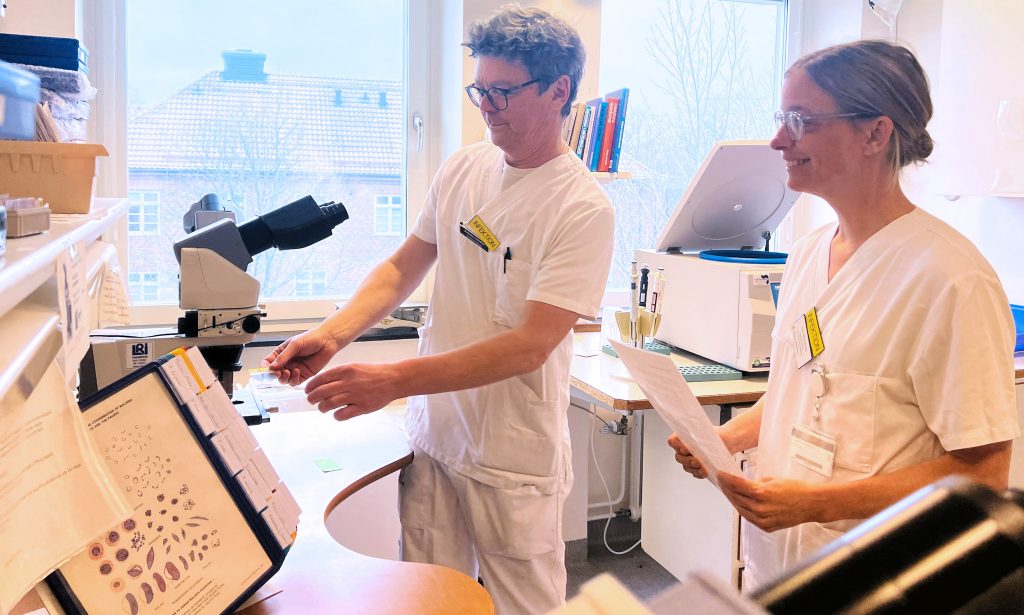 En man och en kvinna klädda i sjukhuskläder framför ett ljusmikroskop