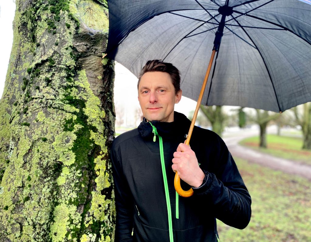 Man i sportjacka lutad mot träd under paraply.