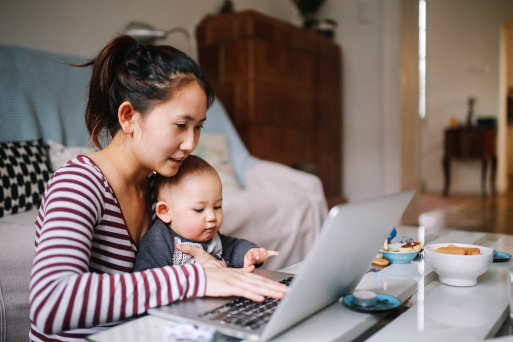 Ung asiatisk kvinna sitter med bäbis i knätt samtidigt som hon har en laptop uppfälld framför sig på bordet och rör tangenterna