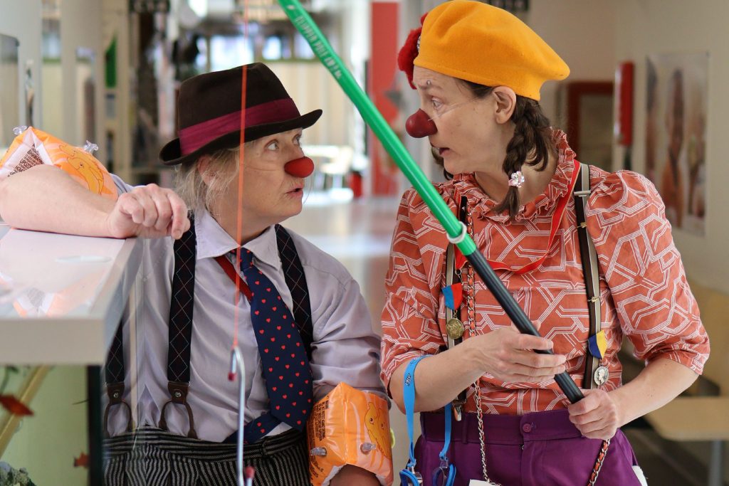 Två clowner i en sjukhuskorridor. Clownen till vänster är Boris, klädd i en hatt, vit skjorta och slips och hängselbyxor. Till höger står Franziz klädd i gulbasker, rödorange blus och lila byxor