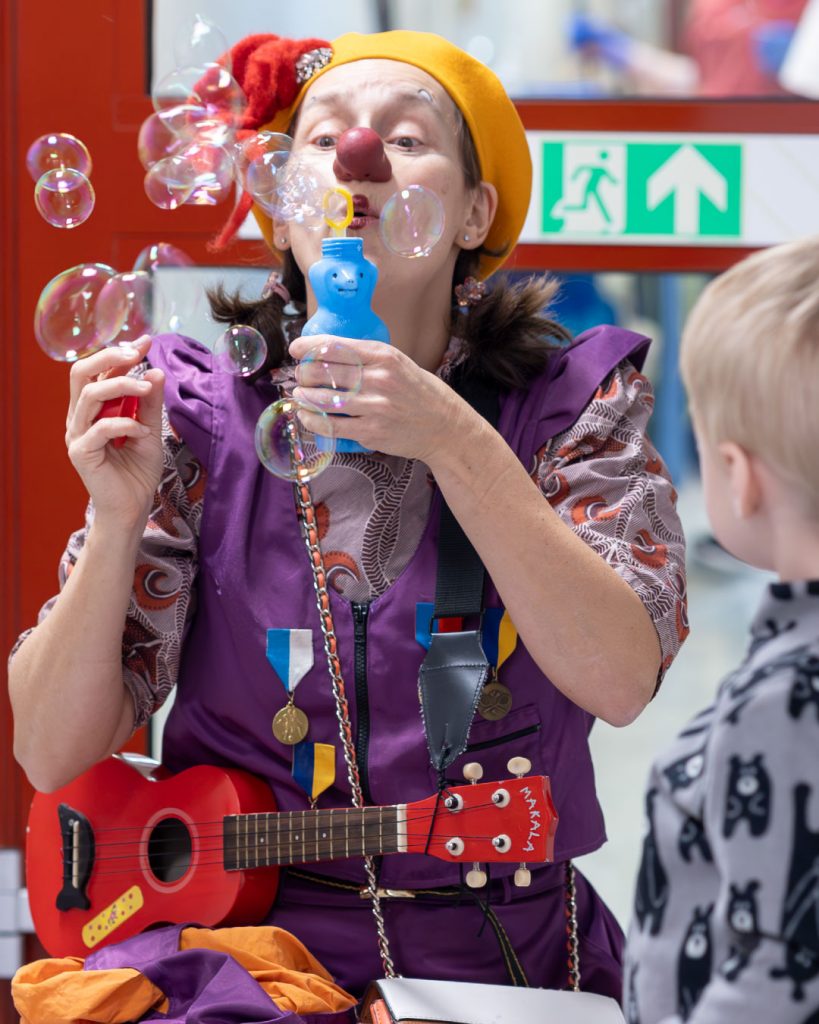 clown röd näsa, gul basker och en ukulele hängande runt halsen blåser såpbubblor. Ett litet barn står bredvid och tittar på