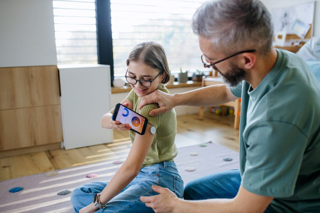 flicka med glukosmätare på armen sitter på golvet och kollar sitt blodsocker mha mobilen. Pappa assisterar