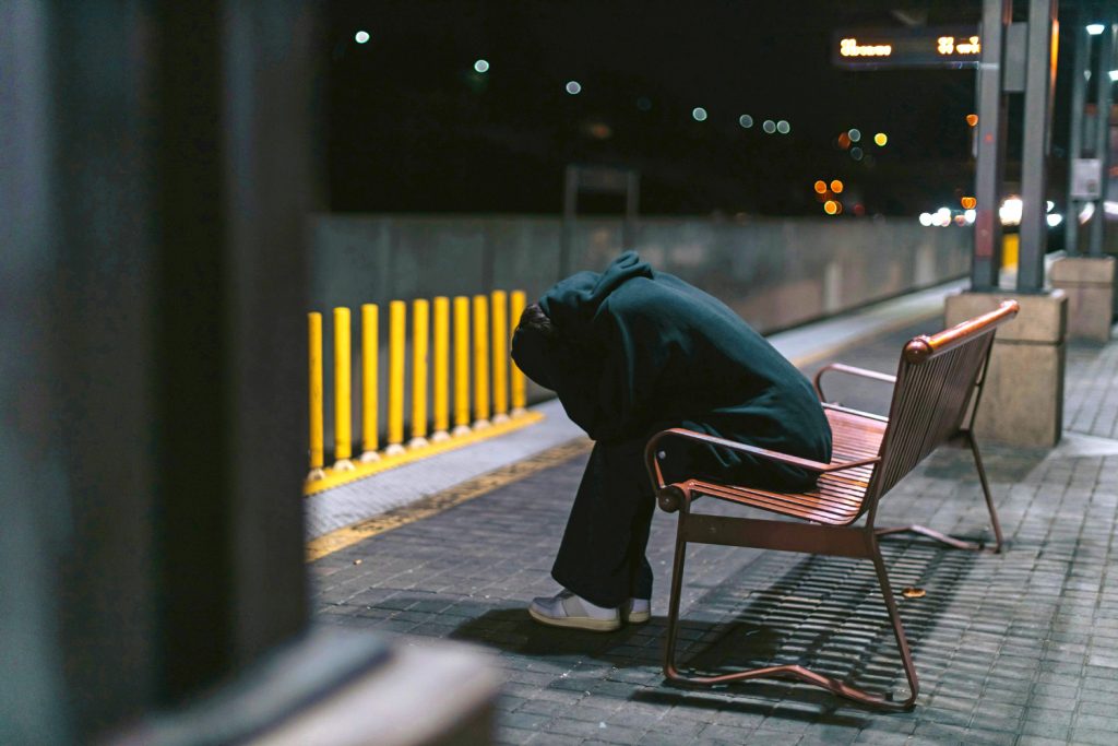 person som sitter på en bänk ute på en station med huvudet i händerna. det är mörkt ute