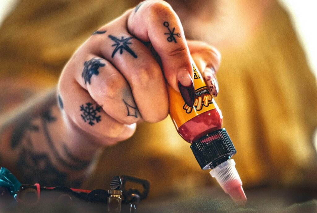 Närbild på tatuerares hand. Det finns svarta tatueringar på varje finger och handen håller i en tub med färg