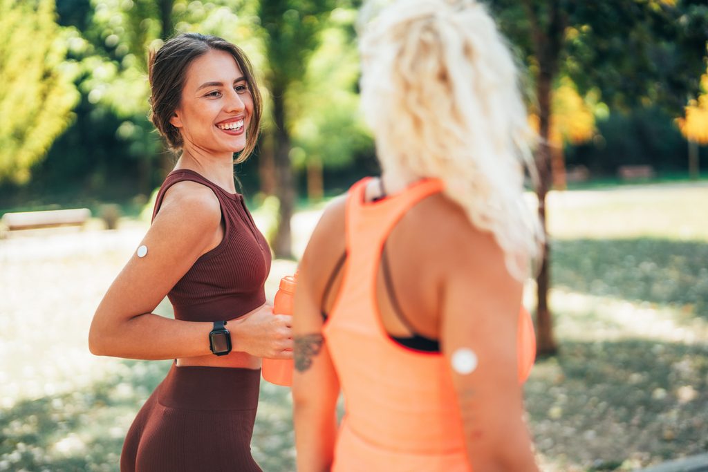 Två unga kvinnor i joggingkläder i en park, båda med glukossensor på armen